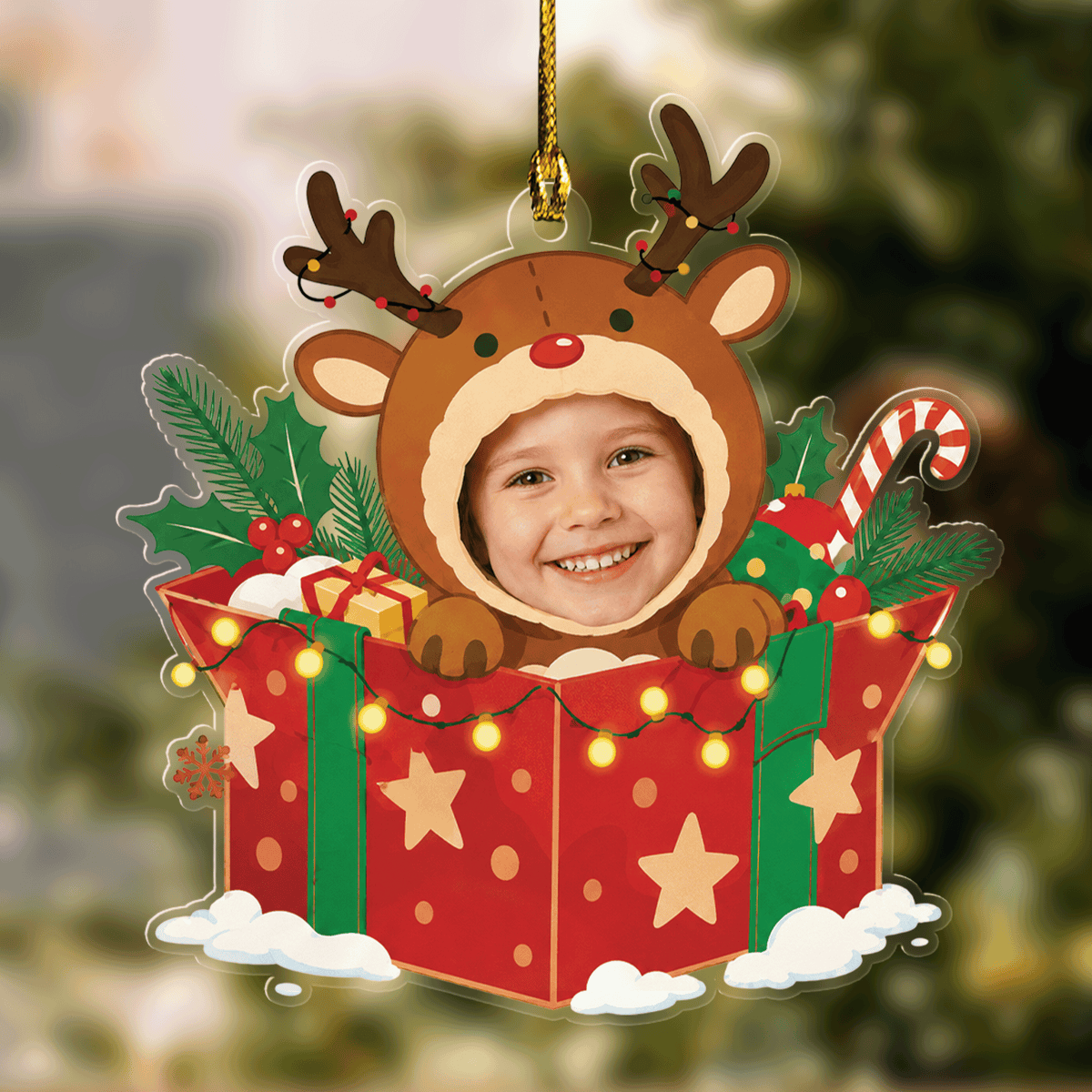 Acrylic ornament of a child's face in a reindeer hood popping out of a red star gift box with candy cane and lights