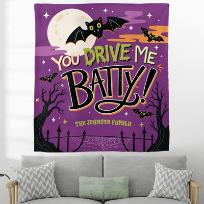 Flat front view of a purple You Drive Me Batty Halloween tapestry with custom family name hanging above a gray sofa