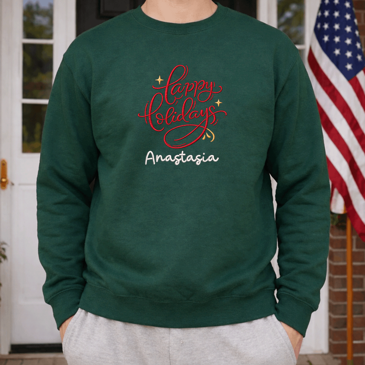 Man wearing a forest green red Happy Holidays name sweatshirt beside an American flag