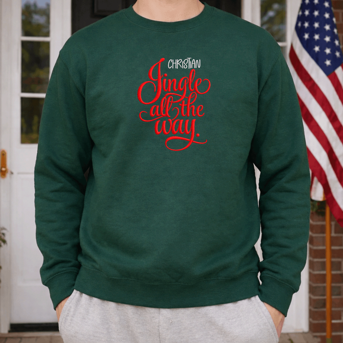 Man wearing a forest green Jingle All the Way sweatshirt beside an American flag