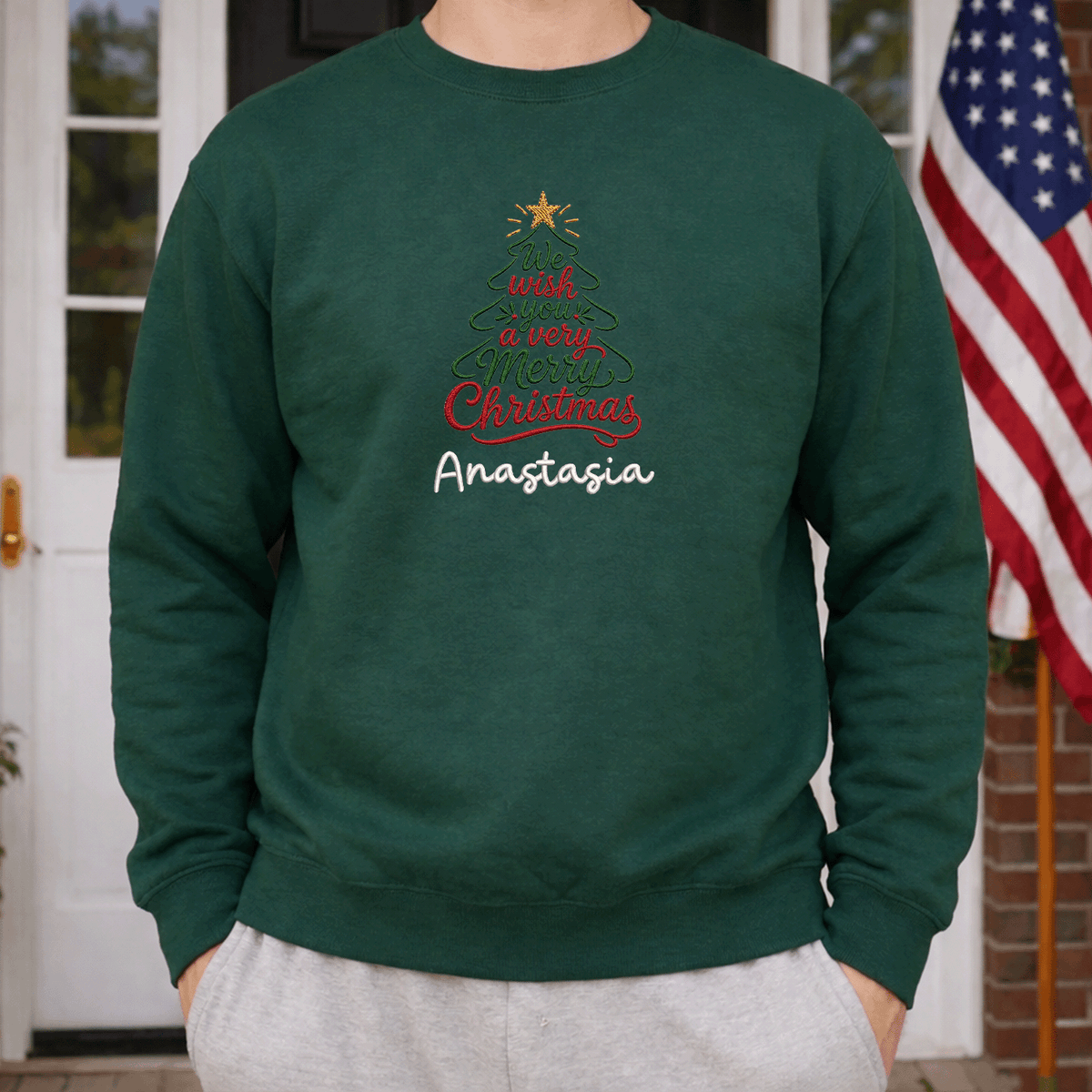 Man wearing a forest green Merry Christmas script tree sweatshirt beside an American flag