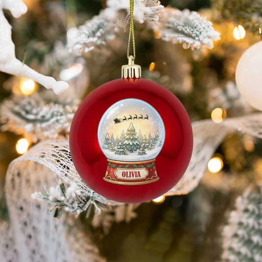 Red ball ornament printed with a snow globe of Santa's sleigh over pine trees and custom name plaque