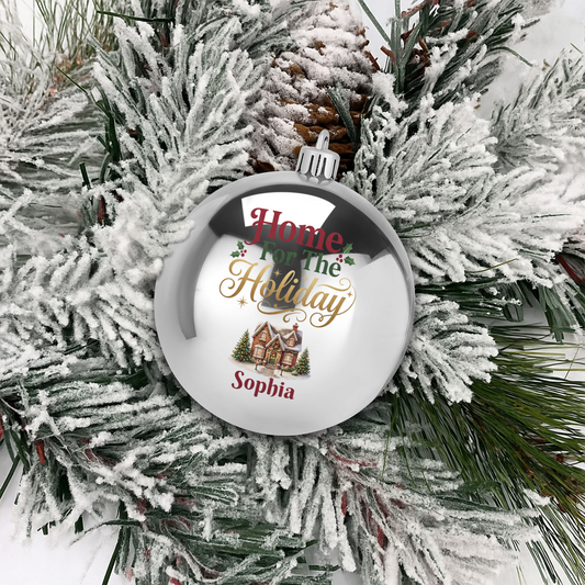 Silver ball ornament printed Home For The Holiday with a snowy winter cottage and custom name on flocked pine