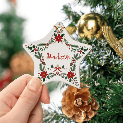 Hand holding the poinsettia wreath ceramic star ornament near a pinecone