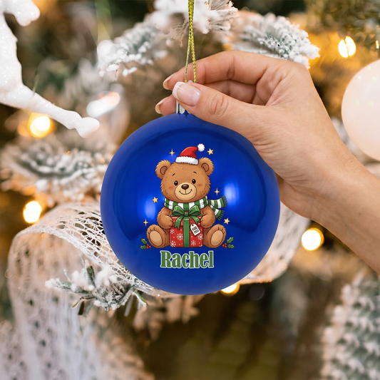 Hand holding a blue Christmas teddy bear name ornament against a blurred tree