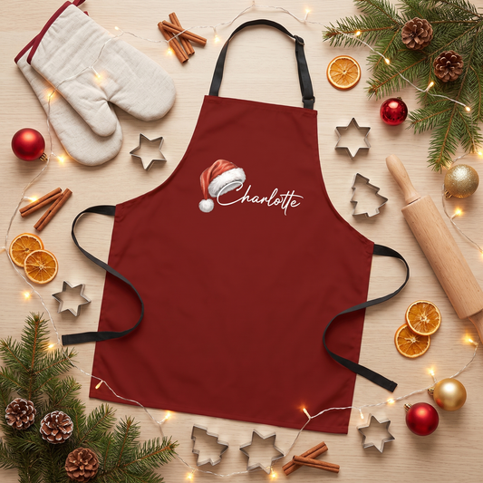 Burgundy apron with a Santa hat on a white script custom name, flat lay with oven mitt and cookie cutters