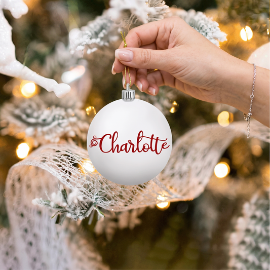 Hand holding a white ball ornament with red script custom name by a Christmas tree