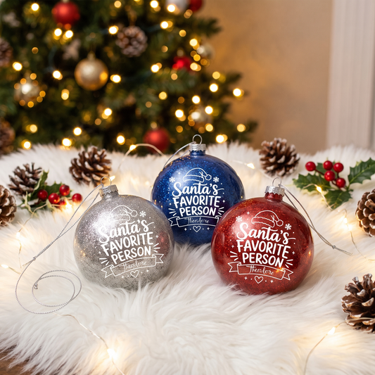 Silver, navy and red Santa's Favorite Person glitter baubles on a white fur mat with pinecones