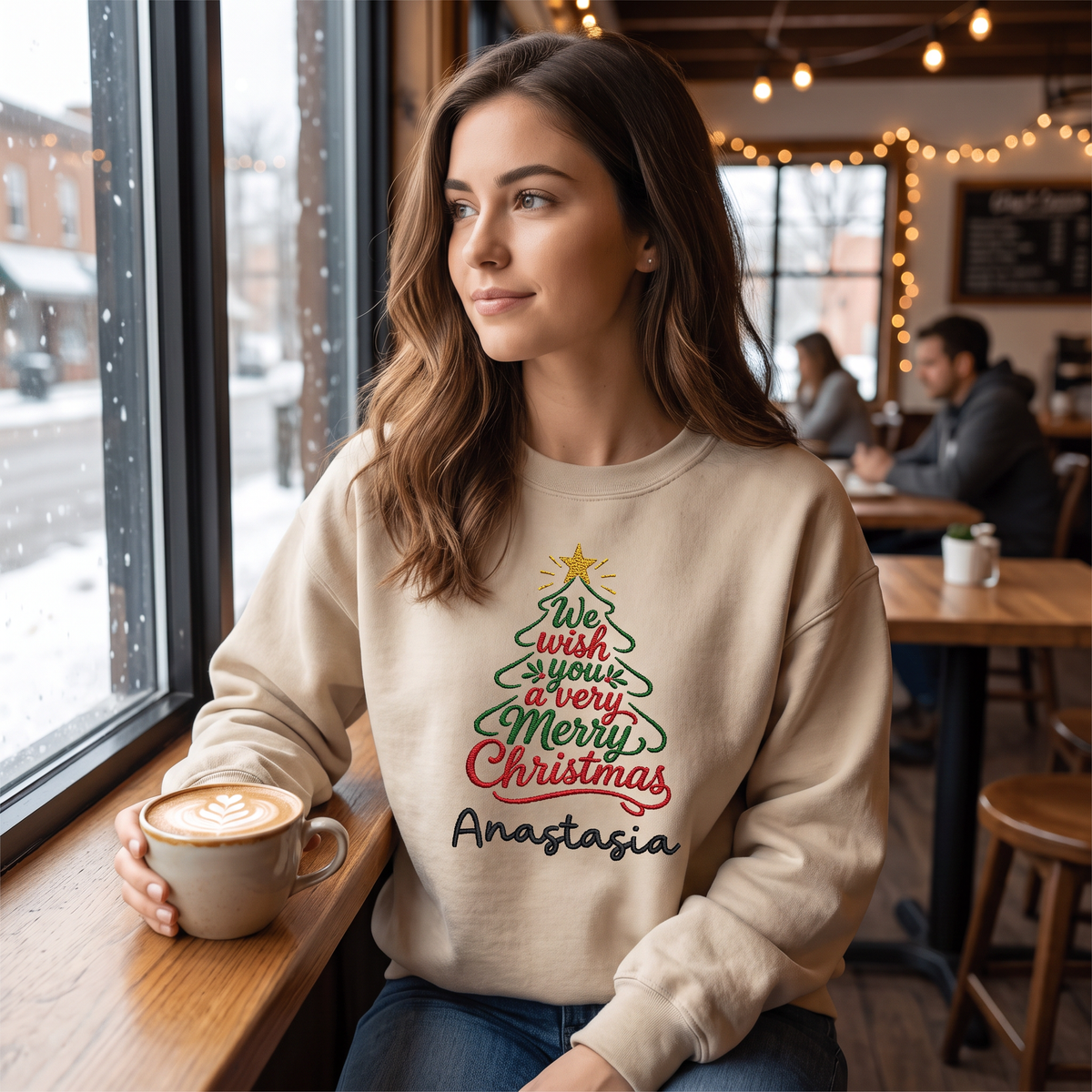 Woman in a sand We Wish You a Merry Christmas sweatshirt holding a latte by a snowy window