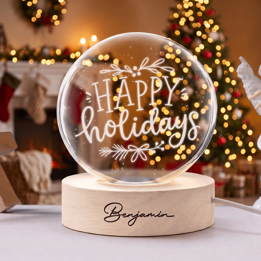 Crystal ball engraved with Happy Holidays calligraphy and sprigs on a wooden base with custom name, Christmas tree behind