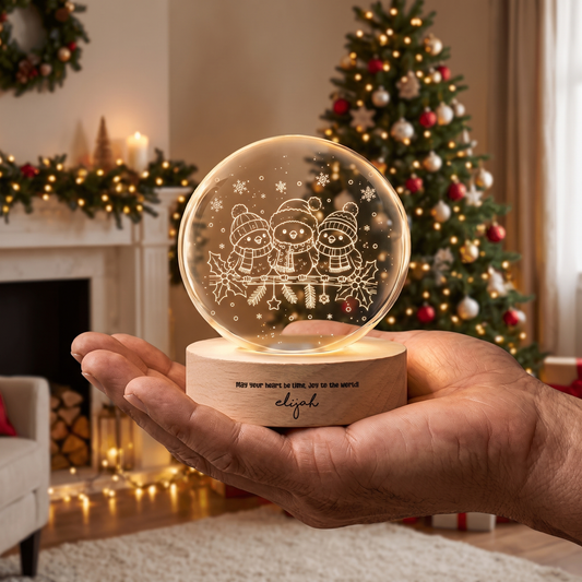 Hand holding a crystal ball engraved with three winter birds in hats and scarves on a holly branch, custom name base