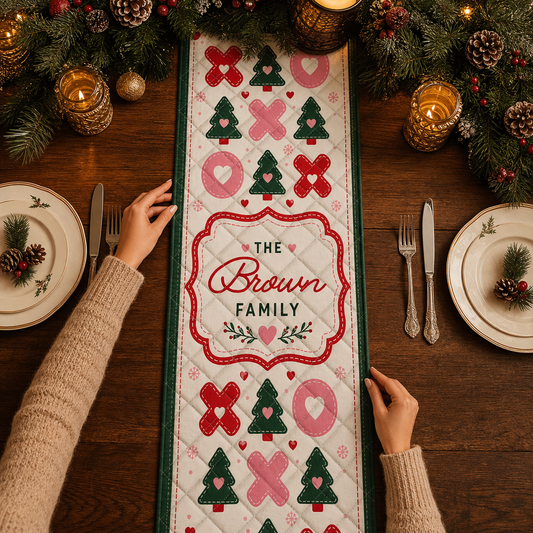 Hands placing a cream quilt runner with pink X and O shapes, green Christmas trees and custom family name frame