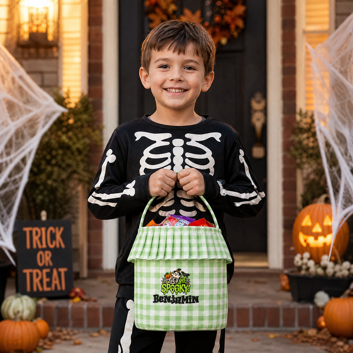 Smiling boy in a skeleton shirt holding a green gingham ruffle trick or treat basket with a custom embroidered name