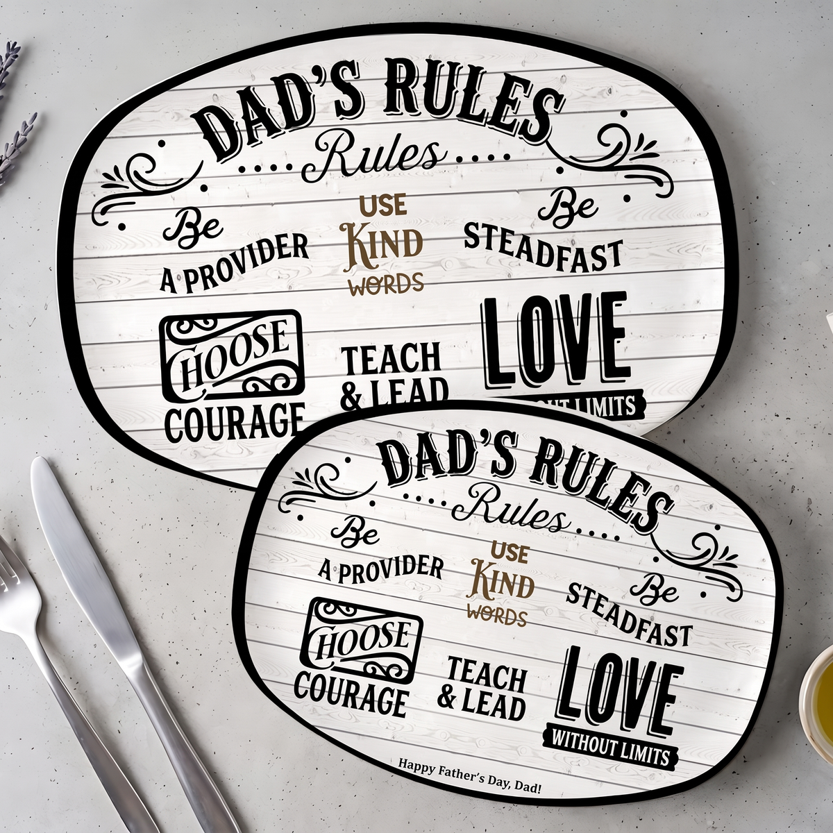 Two sizes of the Dad's Rules oval serving platter overlapping on grey stone beside a knife and fork
