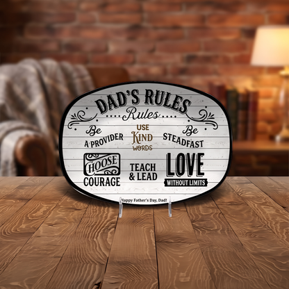 Dad's Rules oval platter displayed upright on a clear stand on a wooden table in front of a brick wall