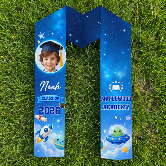 Blue galaxy rocket kid graduation stole with child photo, name Noah, Class of 2026 and Maplewood Academy laid flat on grass