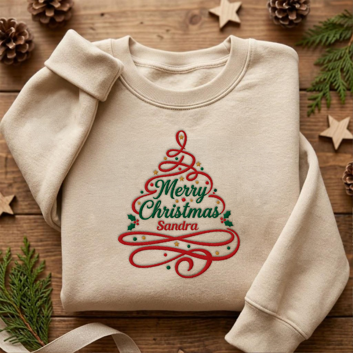 Sand crewneck sweatshirt embroidered with Merry Christmas Ribbon Tree design and a custom name, laid flat with Christmas decor