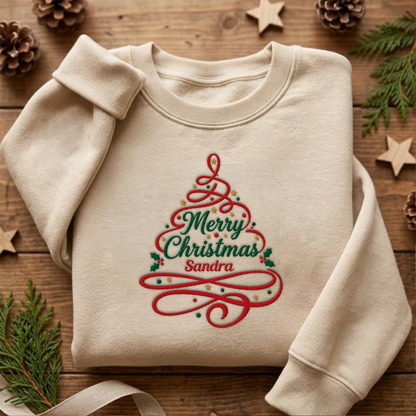 Sand crewneck sweatshirt embroidered with Merry Christmas Ribbon Tree design and a custom name, laid flat with Christmas decor