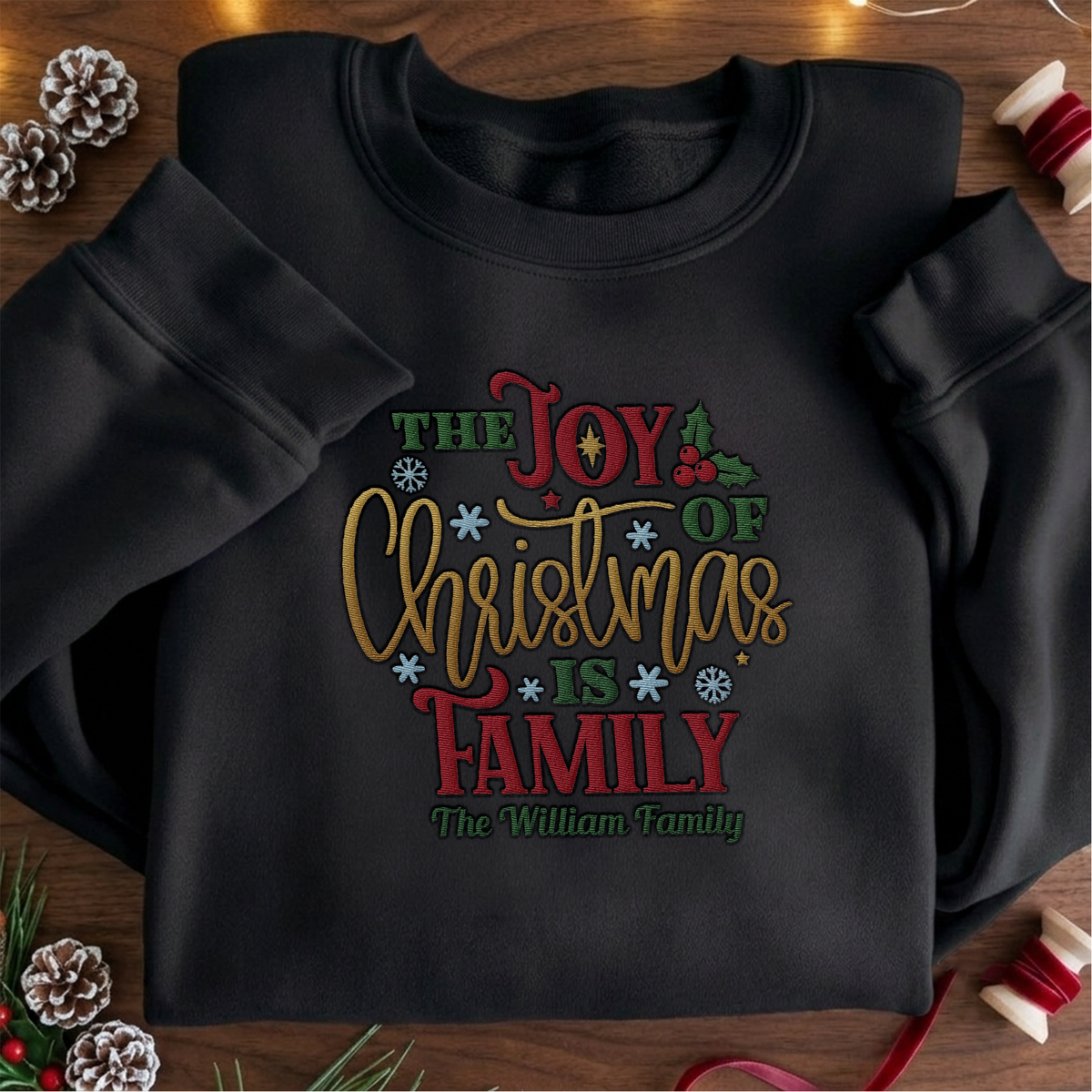 Black crewneck sweatshirt embroidered with The Joy of Christmas Is Family design and a custom family name, laid flat with Christmas decor