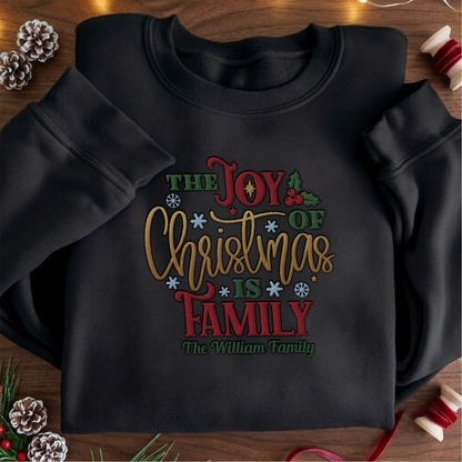 Black crewneck sweatshirt embroidered with The Joy of Christmas Is Family design and a custom family name, laid flat with Christmas decor