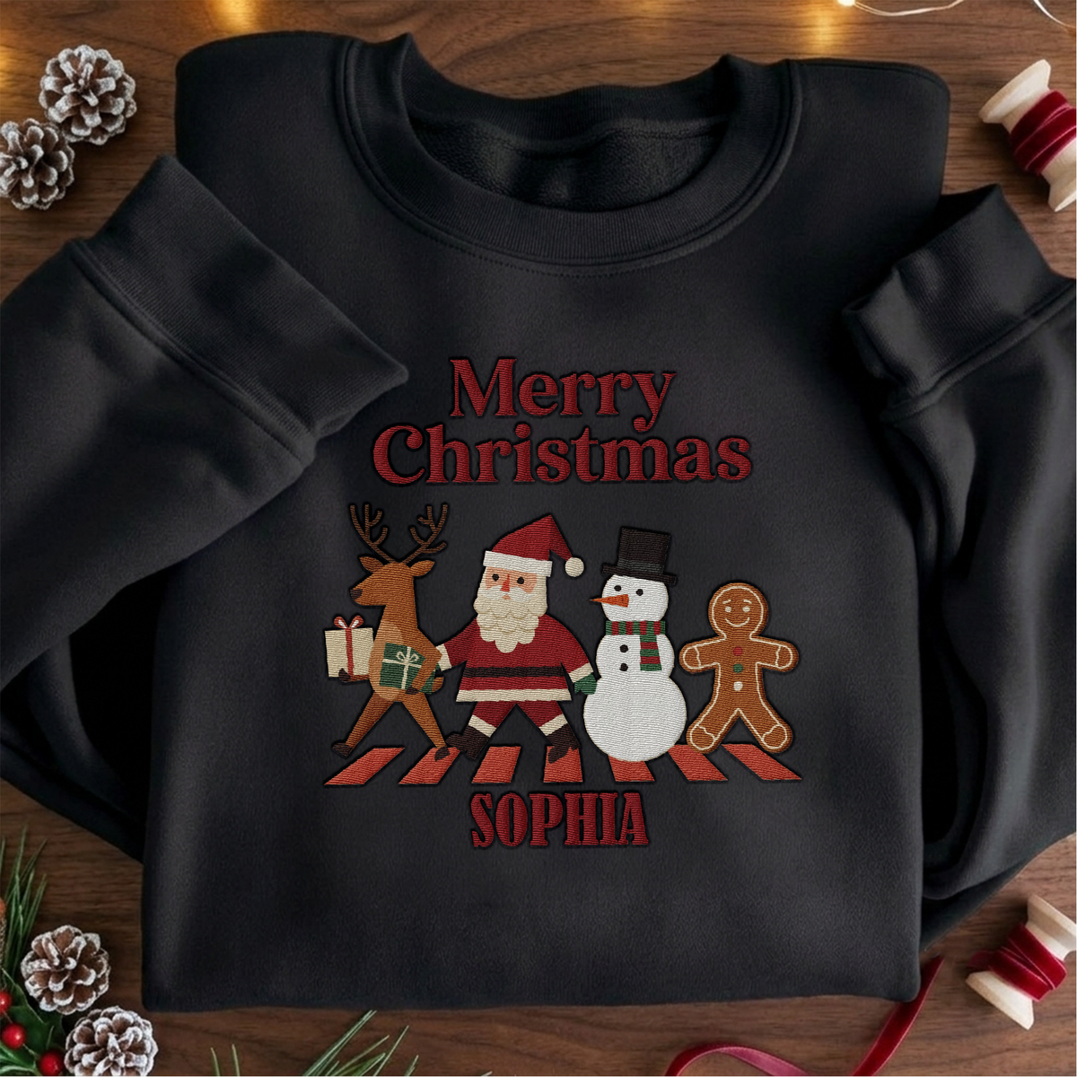 Black crewneck sweatshirt embroidered with Merry Christmas Crew Crossing design and a custom name, laid flat with Christmas decor