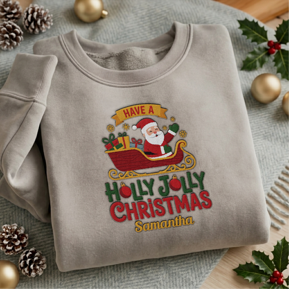 Heather grey crewneck sweatshirt embroidered with Holly Jolly Christmas Santa Sleigh design and a custom name, laid flat with Christmas decor