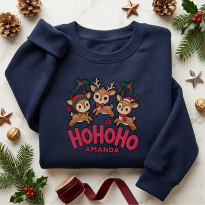 Navy crewneck sweatshirt embroidered with Ho Ho Ho Baby Reindeer design and a custom name, laid flat with Christmas decor