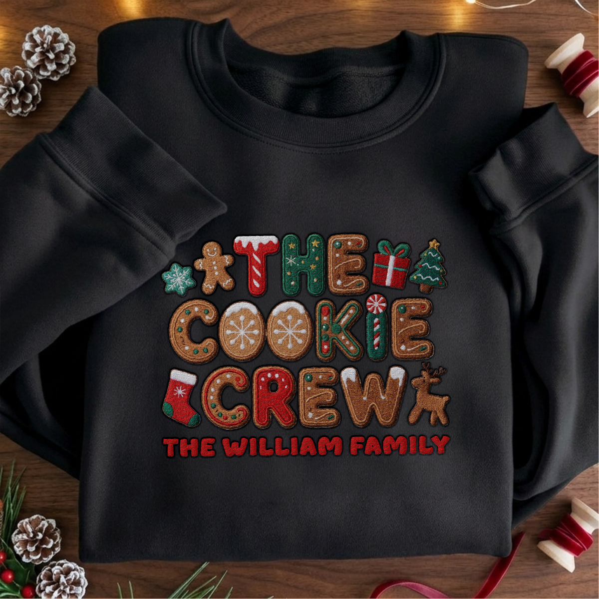 Black crewneck sweatshirt embroidered with The Cookie Crew design and a custom family name, laid flat with Christmas decor